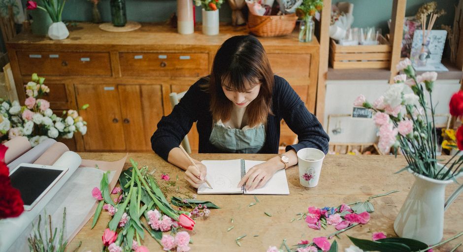 A professional florist with shoulder-length brown hair and wearing a dark cardigan and apron, sits at a wooden worktop in a bright, well-organized flower shop. She is sketching a flower arrangement in