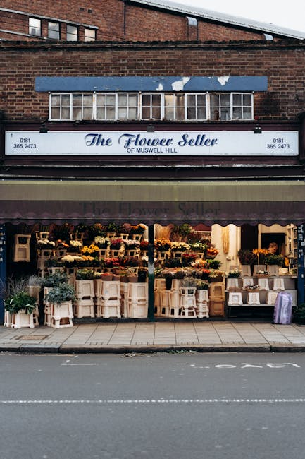 The flower shop 'The Flower Seller of Muswell Hill' features an inviting storefront with an array of fresh floral arrangements displayed on white wooden shelves and stands outside the shop. The displa