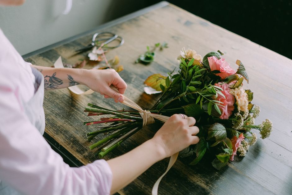 Close-up of a florist's hands wrapping a bouquet of fresh flowers on a wooden table. The bouquet features pink and white roses, greenery, and small white filler flowers. The florist's arms are visible