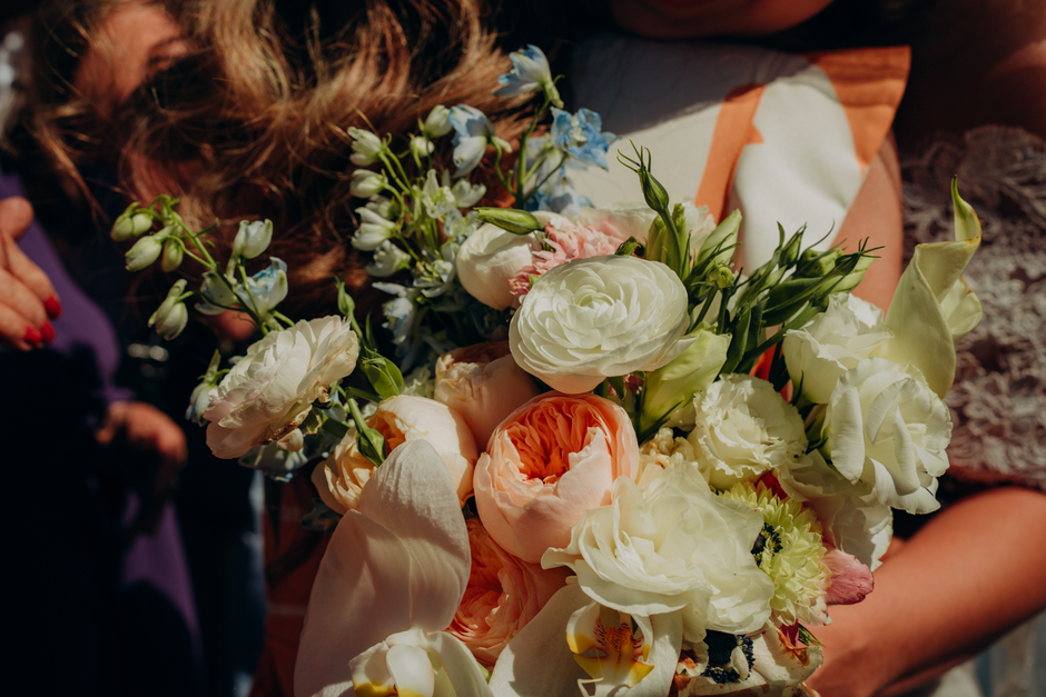 A close-up view of a floral bouquet held by a person, featuring an arrangement of fresh flowers in soft pastel shades of white, cream, peach, and light pink. The bouquet includes large, ruffled roses,