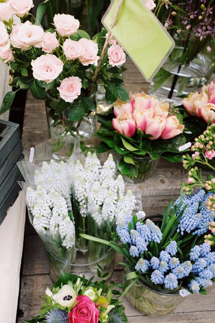 A display of fresh floral arrangements on a wooden surface, including a bouquet of pale pink roses with lush green foliage in a clear glass vase, alongside a cluster of vibrant pink and cream tulips w