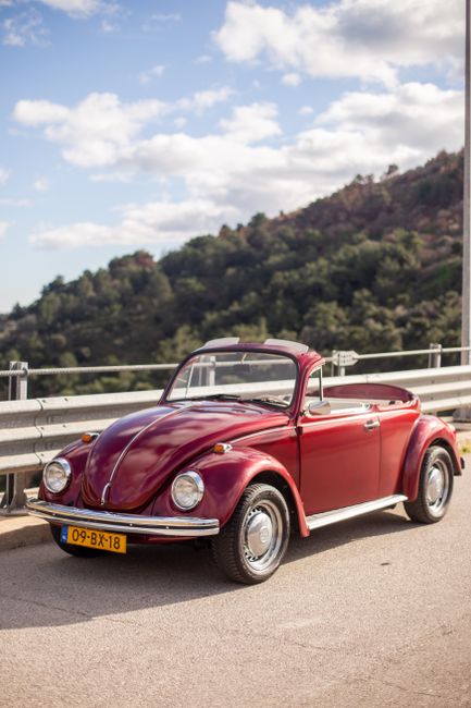 A vintage red Volkswagen Beetle with a convertible top parked on the side of a road, adjacent to a metal guardrail, with a scenic background of green hills and a partly cloudy sky. The car's shiny ext