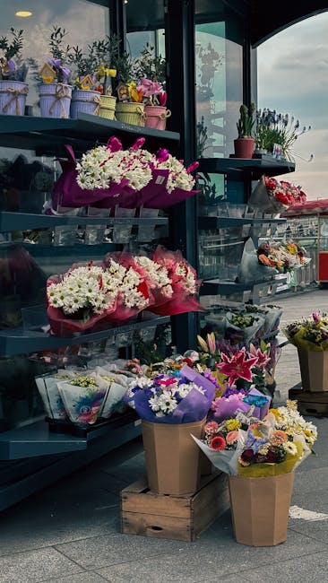 A display of various fresh floral arrangements at Gipsy Hill Florist, showcasing bouquets wrapped in colorful tissue paper and placed on a metal shelving unit inside a glass-fronted shop. The arrangem