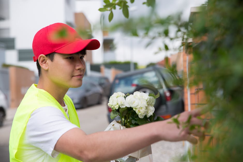 A young delivery person wearing a red cap and a neon yellow vest is seen placing a floral gift, consisting of white hydrangeas, into a green bush in an outdoor setting. The arrangement is held with bo